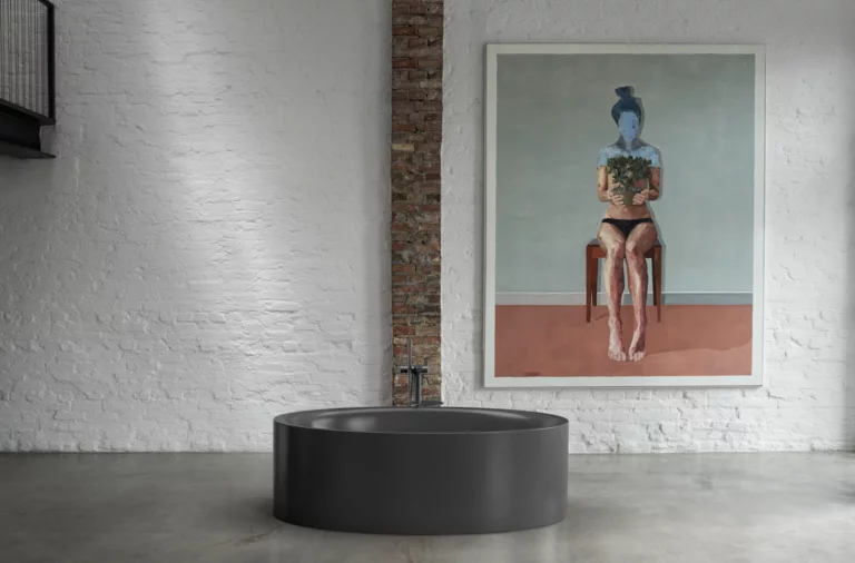 Freistehende, ovale Badewanne in Schwarz vor weißer Backsteinwand mit Kunstwerk und freigelegtem Mauerstreifen.